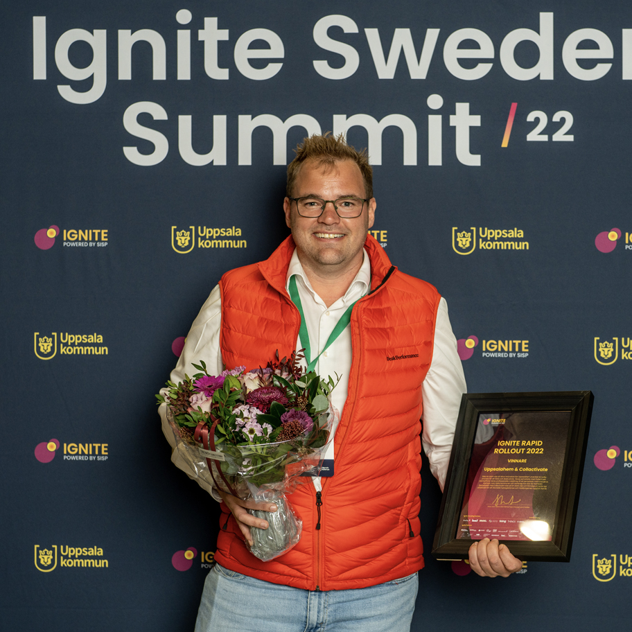 En man med glasögon och kort hår står framför en backdrop med texten "Ignite Sweden Summit /22". Han bär en vit skjorta och en orange väst och har en grön namnbricka runt halsen. Han håller en bukett blommor i ena handen och en inramad utmärkelse i den andra, där det står "IGNITE RAPID ROLLOUT 2022" och "VINNARE Uppsteam & Collectivate". Han ler mot kameran.