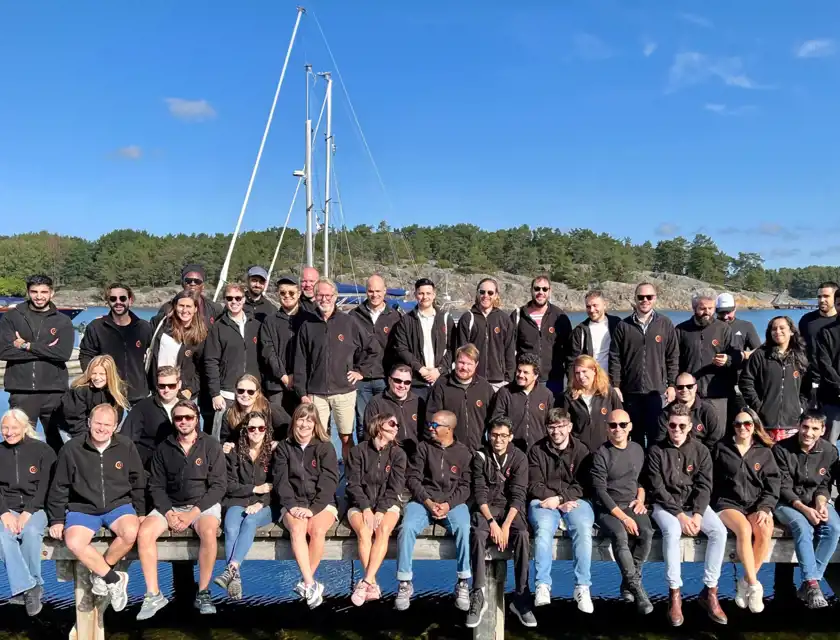 En stor grupp människor poserar tillsammans på en brygga vid vattnet, omgiven av skärgårdsmiljö med båtar och grönskande öar i bakgrunden. De flesta i gruppen bär svarta jackor med en logotyp på bröstet. De sitter och står avslappnat, många med leenden, medan solen lyser över scenen.