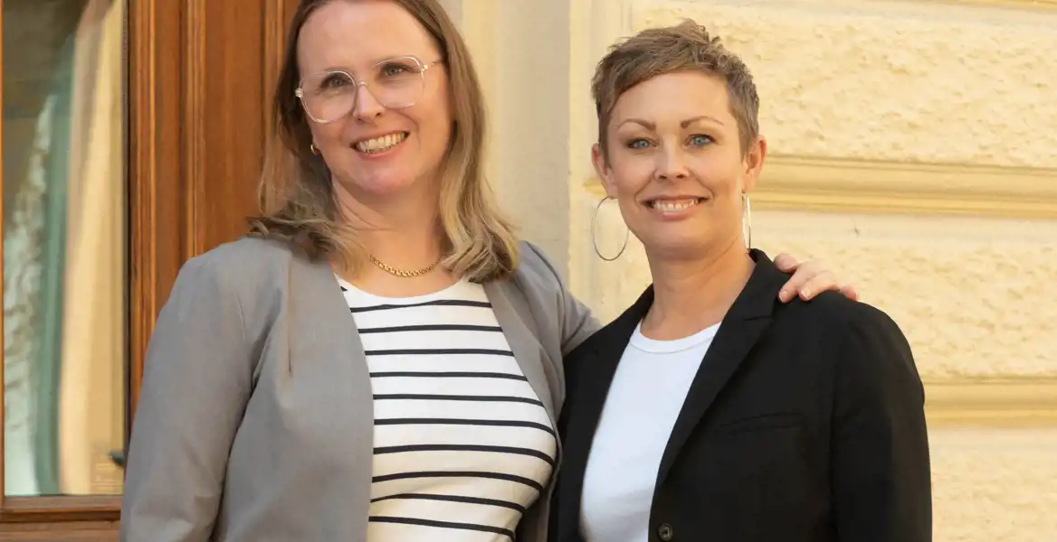 Två kvinnor står tillsammans utomhus framför en byggnad med en trädetaljerad dörr och en ljus fasad. De ler och har en avslappnad men professionell hållning. Kvinnan till vänster bär en grå kostym med en vit- och svartrandig topp, glasögon och en guldkedja. Kvinnan till höger har kort hår och bär en svart kavaj över en vit t-shirt, samt stora ringörhängen. De ser ut att vara kollegor eller samarbetspartners.