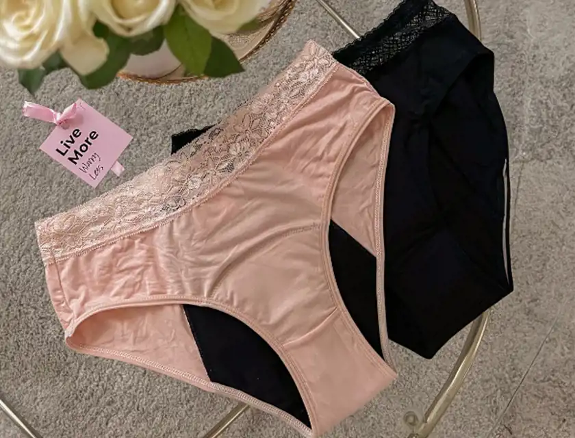 Två par menstruationsunderkläder ligger på ett glasbord, ett i ljusrosa och ett i svart. Båda har spetsdetaljer i midjan och en absorberande svart insida. Ett rosa etikettkort med texten "Live More" och "Happy" sitter fast i det ljusrosa paret. I bakgrunden syns en vas med vita rosor och en ljus matta.