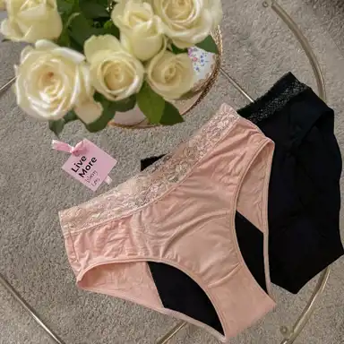 Två par menstruationsunderkläder ligger på ett glasbord, ett i ljusrosa och ett i svart. Båda har spetsdetaljer i midjan och en absorberande svart insida. Ett rosa etikettkort med texten "Live More" och "Happy" sitter fast i det ljusrosa paret. I bakgrunden syns en vas med vita rosor och en ljus matta.