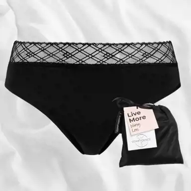 Ett par svarta menstruationsunderkläder med en elegant spetskant ligger på ett vitt tyg. Bredvid dem finns en svart förvaringspåse med en rosa etikett som har texten "Live More Worry Less" och en mindre lapp med logotypen "COMFYDENCE". Underkläderna har en diskret tygetikett i sidan med varumärkets namn.