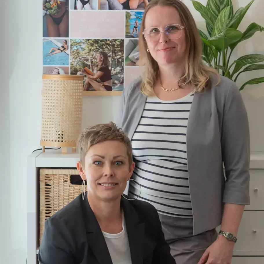 Två kvinnor står tillsammans utomhus framför en byggnad med en trädetaljerad dörr och en ljus fasad. De ler och har en avslappnad men professionell hållning. Kvinnan till vänster bär en grå kostym med en vit- och svartrandig topp, glasögon och en guldkedja. Kvinnan till höger har kort hår och bär en svart kavaj över en vit t-shirt, samt stora ringörhängen. De ser ut att vara kollegor eller samarbetspartners.