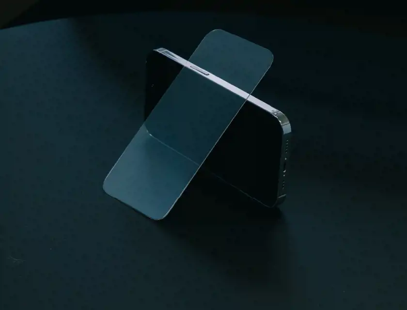Ett skärmskydd balanserar lutat mot en smartphone som står upprätt på ett mörkt bord. Telefonens skärm är svart och reflekterar svagt ljuset i rummet. Skärmskyddet är genomskinligt och har rundade kanter, vilket visar dess tunna och exakta passform för enheten. Bilden har en stilren och minimalistisk känsla med en dämpad ljussättning.