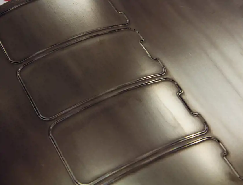 En närbild av en metallplåt med utskurna konturer av skärmskydd. De tunna, precisionsskurna linjerna formar rektangulära former med rundade hörn och små utskärningar, vilket antyder att detta är en del av tillverkningsprocessen för skärmskydd till smartphones. Plåtens yta har en borstad metallfinish, och ljuset reflekteras mjukt över materialet, vilket skapar en industriell och teknisk känsla.