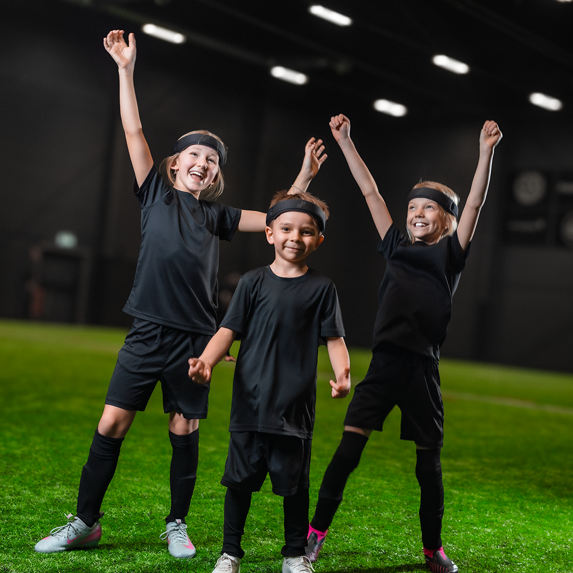 Tre barn i fotbollskläder bär huvudskydd från DAJE Prevent och står på en fotbollsplan med armarna i luften.
