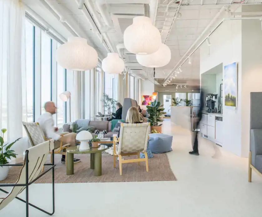 En ljus och modern kontorsmiljö med stora fönster, skandinavisk design och gröna växter. Mjuka sittgrupper, stilrena möbler och papperslampor skapar en inbjudande atmosfär. Människor arbetar och umgås i en avslappnad miljö.