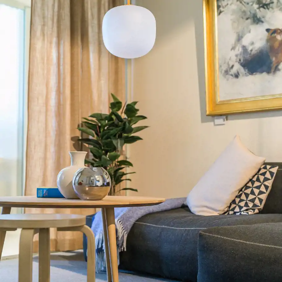 Bilden visar ett stilrent och inbjudande rum med en modern och harmonisk inredning. En mjuk soffa med dekorativa kuddar och en filt skapar en avslappnad atmosfär. Ett runt träbord i förgrunden är dekorerat med eleganta vaser och en bok. En lampa med en enkel, rund design hänger från taket och ger ett mjukt ljus. En stor tavla med en guldram pryder väggen, och en grön växt i bakgrunden bidrar till en naturnära känsla. Ljusa gardiner och ett fönster släpper in naturligt ljus, vilket förstärker rummets varma och ombonade känsla.