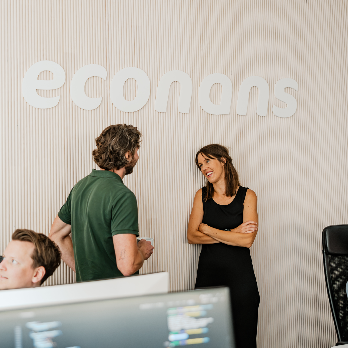 En man i grön pikétröja och en kvinna i svart klänning står och samtalar leende framför en vägg med texten 'econans'. I förgrunden syns två datorskärmar och en person som sitter vid en arbetsstation.
