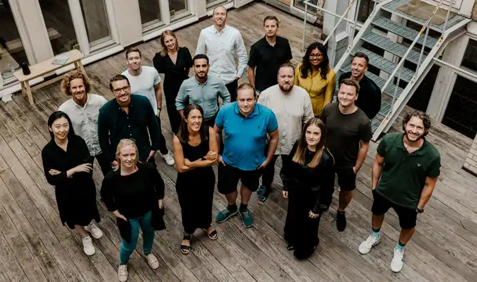 En grupp på 17 personer står tillsammans på en träaltan och ser upp mot kameran med glada uttryck. De är klädda i en mix av avslappnade och affärsmässiga kläder. I bakgrunden syns en utomhustrappa, stora fönster och en liten träbänk med några böcker.