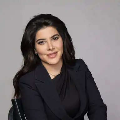 Bilden visar en kvinna med långt, mörkt hår och en professionell klädstil i svart. Hon har ett vänligt uttryck och bär en diskret guldkedja. Bakgrunden är neutral och ljus, vilket ger en stilren och professionell känsla.