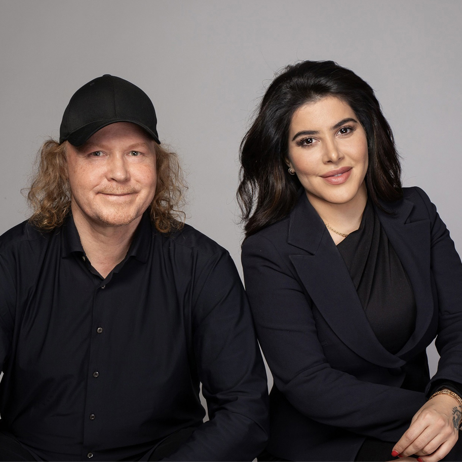 Bilden visar två personer i mörka kläder mot en neutral bakgrund. Mannen har axellångt hår, keps och en avslappnad hållning. Kvinnan har långt, mörkt hår och en professionell klädstil. Båda har ett vänligt uttryck.