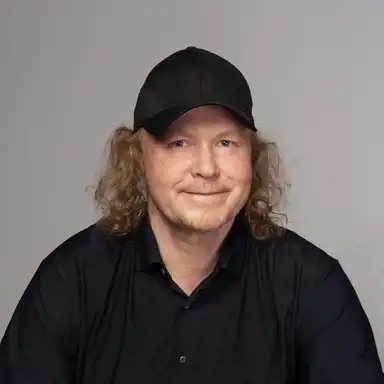 En man med långt lockigt hår och en svart keps poserar för ett porträtt. Han har ett vänligt uttryck och bär en svart skjorta. Bakgrunden är neutral och ljusgrå, vilket ger bilden ett professionellt och stilrent intryck.