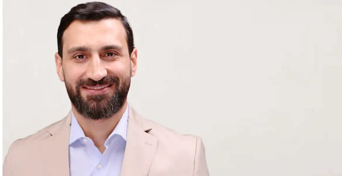 Porträtt av en leende man med mörkt hår och skägg, klädd i en ljusbeige kavaj och ljusblå skjorta. Han har en varm och vänlig utstrålning, och bakgrunden är ljus och neutral.