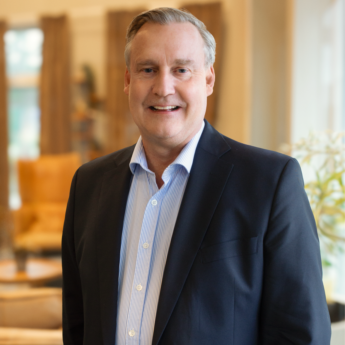 En man i kostym står inomhus i en ljus och varm miljö. Han ler mot kameran och bakgrunden visar soffor, gardiner och mjuka färger som ger ett välkomnande intryck.