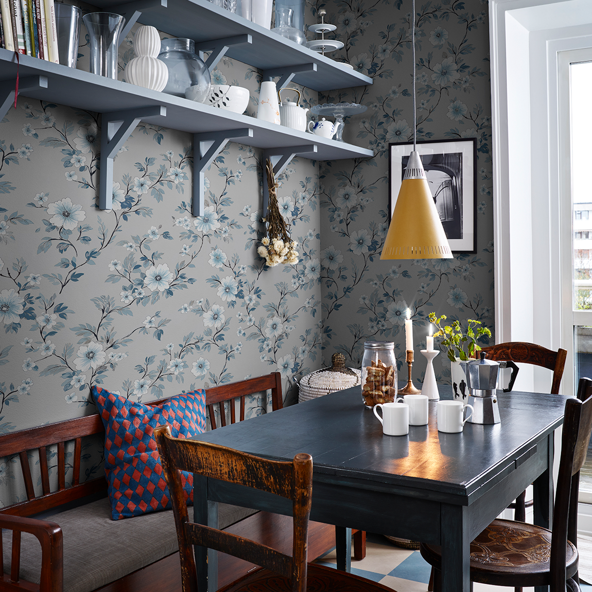 Ett kök med blommig tapet i blå toner från Familjetapeter. Ett mörkt träbord står mitt i rummet med vita muggar, en espressokanna och tända ljus. Runt bordet finns äldre trästolar och en bänk med en färgglad kudde. Ovanför hänger öppna hyllor med glas och porslin samt en gul taklampa. Ljuset faller in från ett fönster till höger.