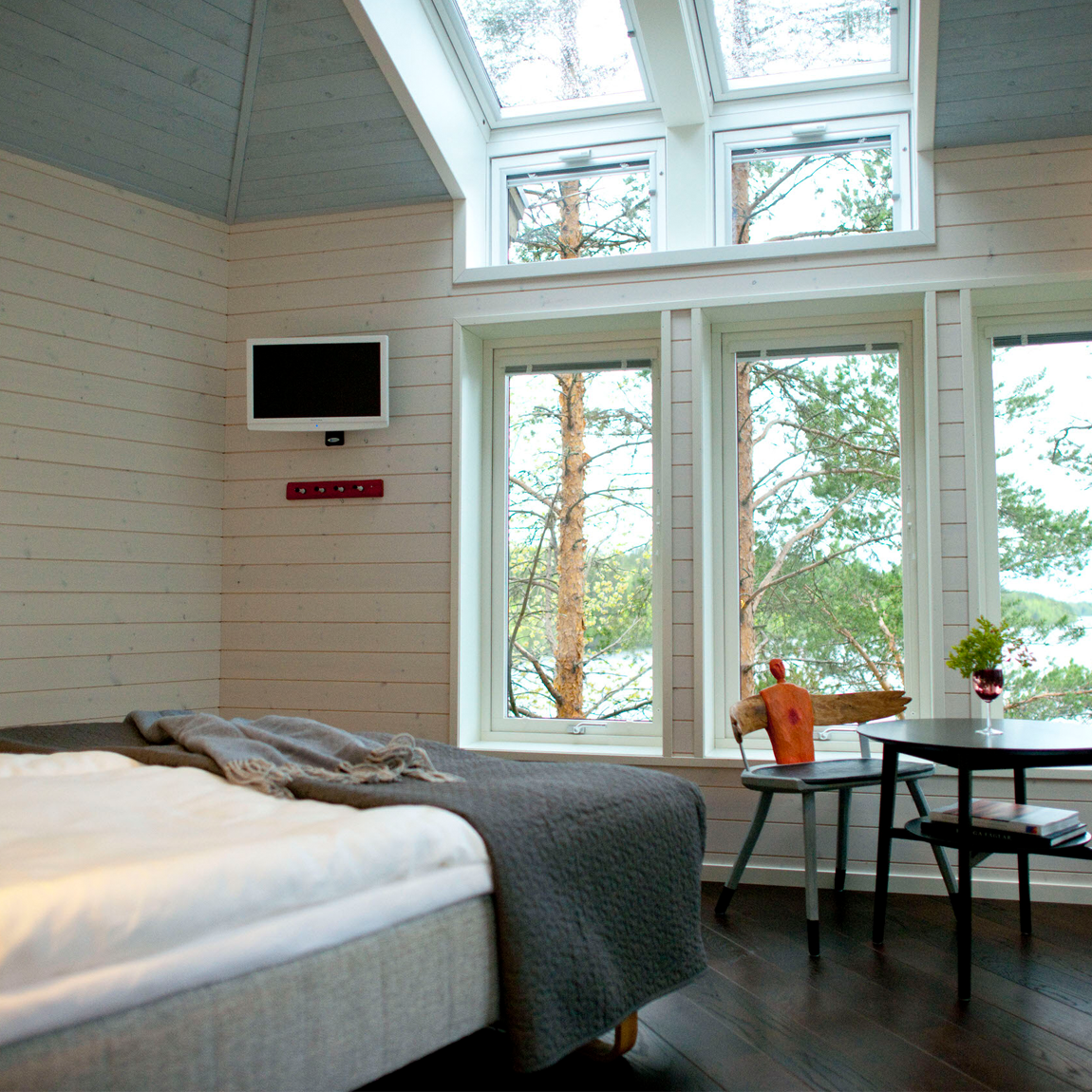 Ett ljust sovrum med stora fönster mot tallar och sjöutsikt. Rummet har en bäddad säng, ett litet bord med två trästolar och takfönster som släpper in mycket dagsljus.