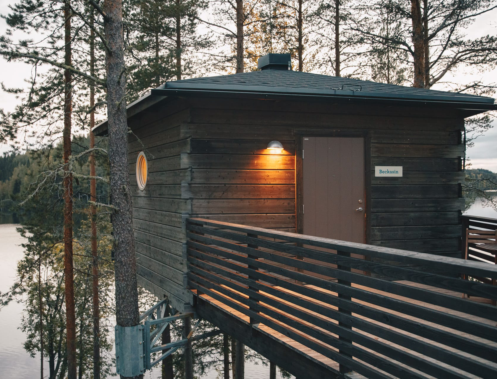 En liten trähytt från Granö Beckasin står upphöjd bland tallar med utsikt över en sjö. En ramp med mörkt räcke leder fram till dörren, och en varm utelampa lyser på fasaden i skymningen.