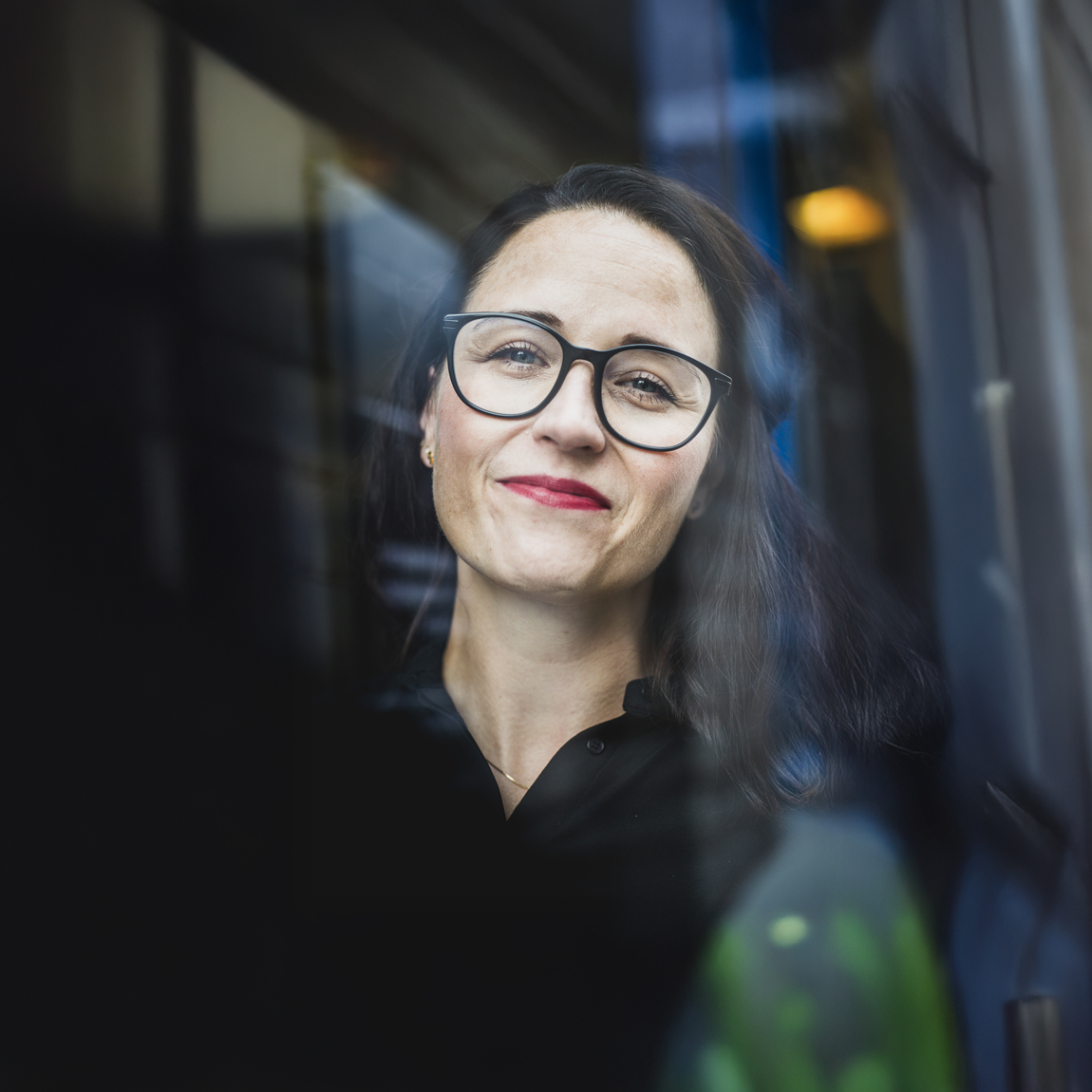 Ett porträtt av en kvinna med långt mörkt hår och glasögon som står inomhus och ler milt mot kameran. Bilden är tagen genom en glasruta som skapar mjuka reflektioner runt motivet.
