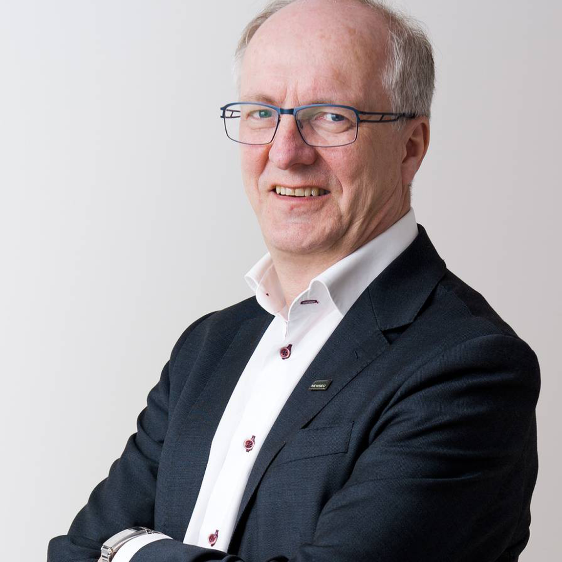 Ett porträtt av en man i kostym som står med armarna i kors och ler mot kameran, mot en ljus bakgrund.