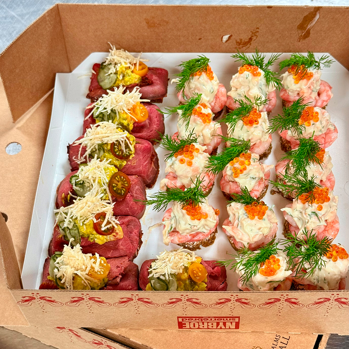 En öppen takeaway-låda med små smörgåsar toppade med rostbiff, räkröra, dill och rom, upplagda i prydliga rader.