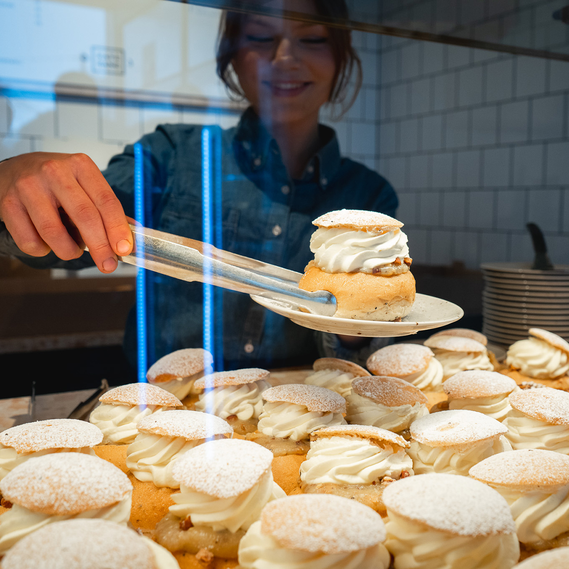 En person bakom en disk lägger upp en semla med grädde och florsocker bland flera semlor i ett kafé.