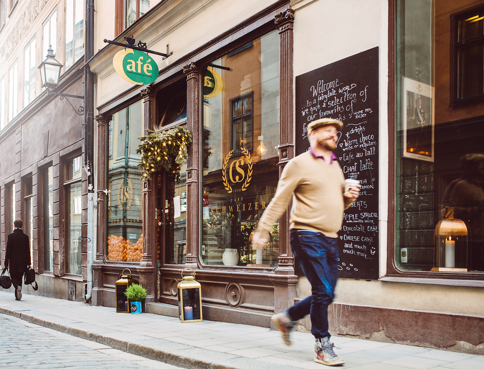 Gatubild av Cafe Schweizers fasad med skyltfönster och uteskylt, där en person går förbi på trottoaren utanför kaféet.