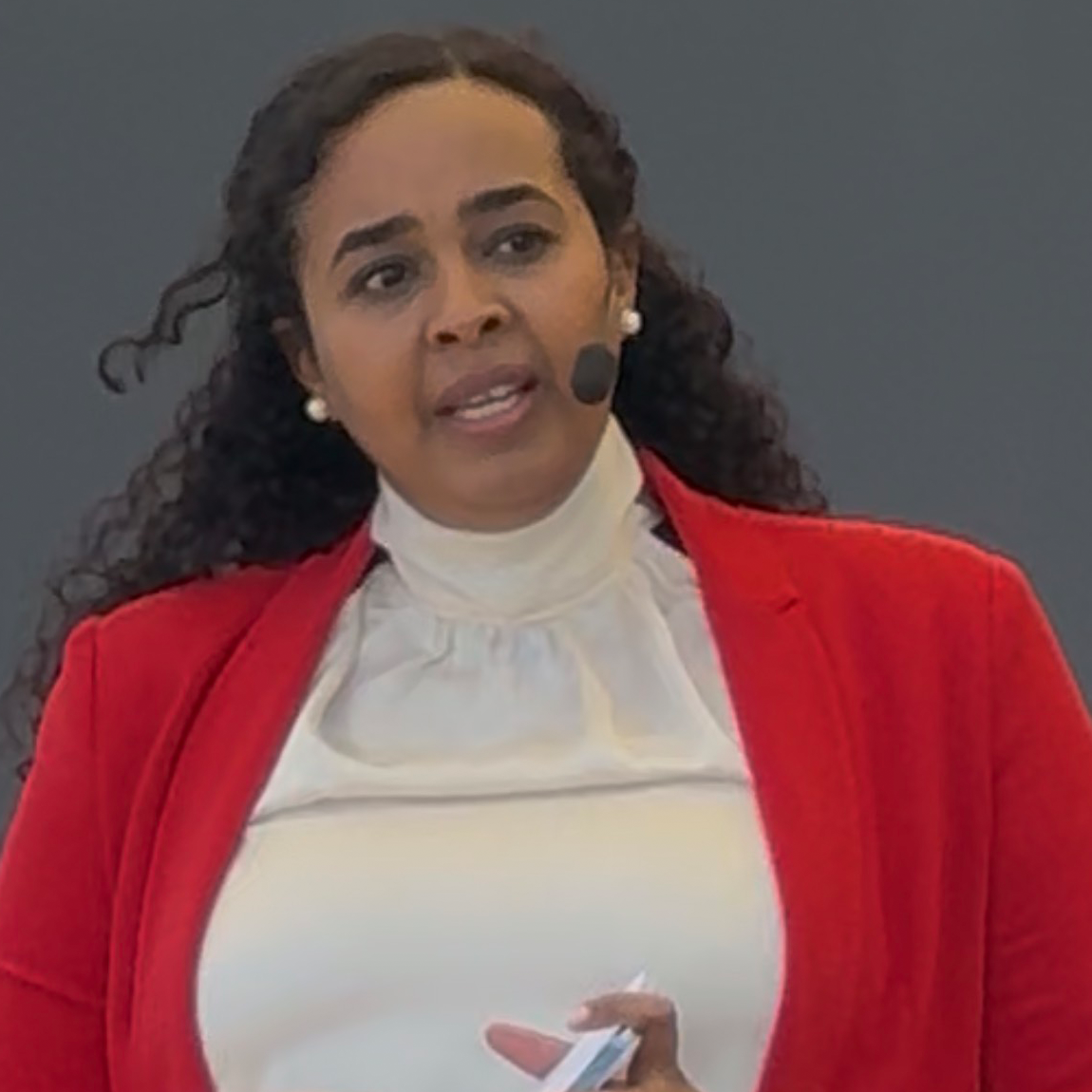 En kvinna håller en presentation och bär en röd kavaj och ljus blus. Hon har ett headset med mikrofon och står framför en neutral bakgrund.