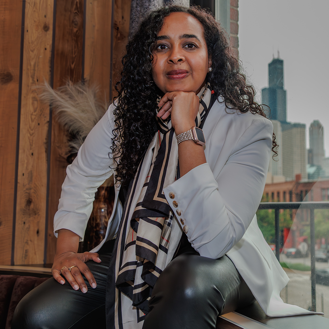 En kvinna sitter avslappnat inomhus med ena handen lutad mot hakan. Hon bär en ljus kavaj, mönstrad scarf och mörka byxor. Bakgrunden visar träpaneler och ett fönster med utsikt över en stadssilhuett.