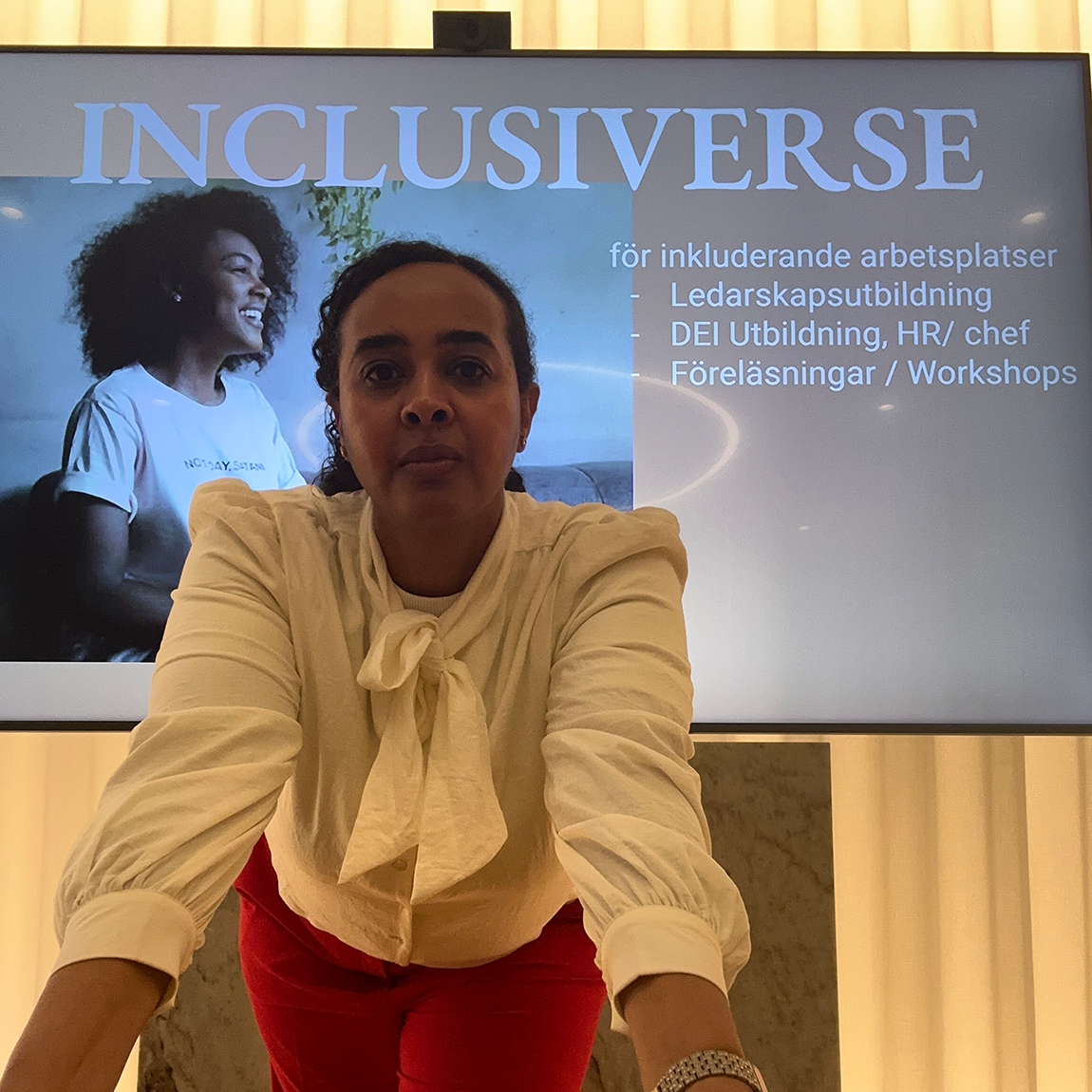 En person står framför en stor skärm som visar texten ”Inclusiverse” och information om utbildningar och workshops för inkluderande arbetsplatser. Personen bär en ljus blus med knut och röda byxor och lutar sig lätt framåt mot kameran. Skärmen bakom visar en leende person och listade tjänster.