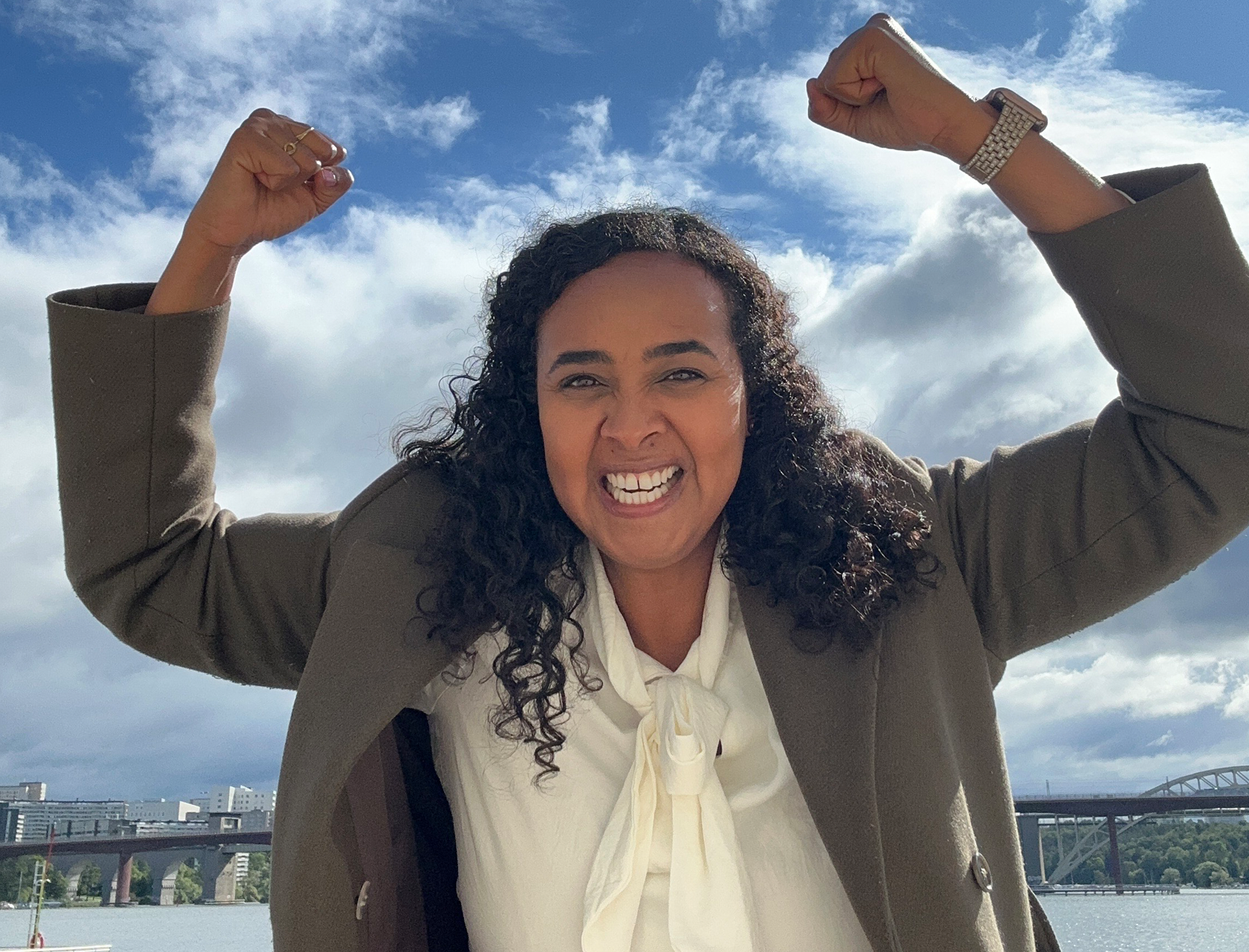 En kvinna står utomhus vid vattnet och lyfter armarna i en segergest. Hon ler stort och bär en ljus blus och mörk kappa. I bakgrunden syns en bro, vatten och bebyggelse under en himmel med moln och blå partier.