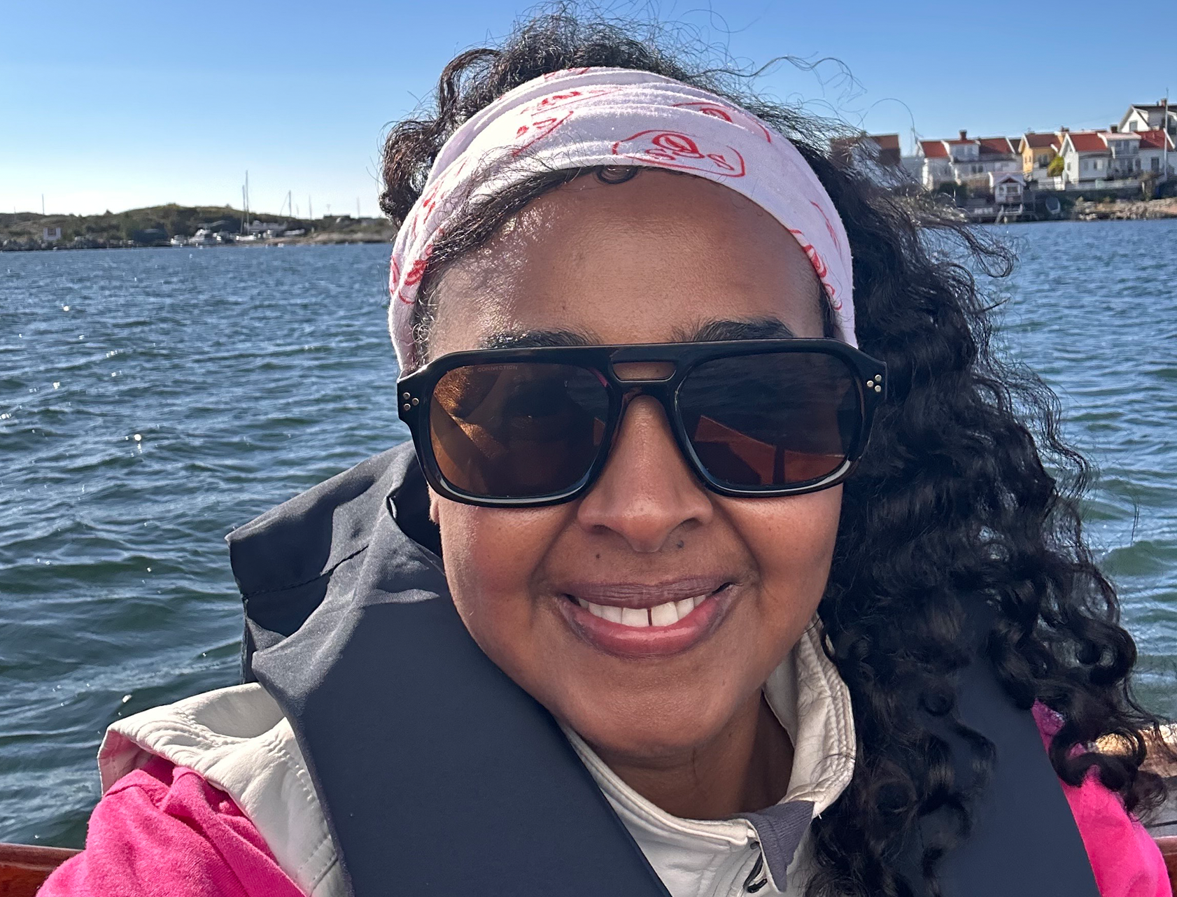 En person tar en selfie på en båt en solig dag. Hen bär solglasögon, ett mönstrat pannband och flytväst. I bakgrunden syns havet, småbåtar och hus längs kusten.