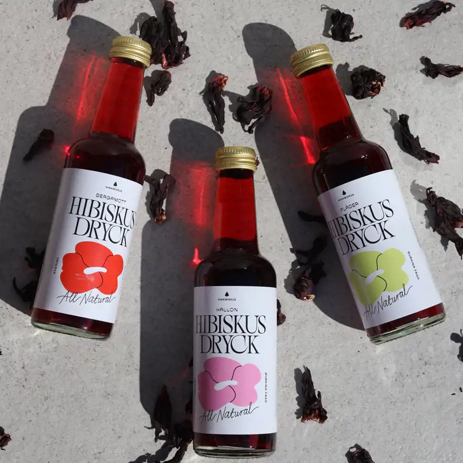 Tre flaskor Hibiskus Dryck ligger på en ljus yta, omgivna av torkade hibiskusblommor. Varje flaska har en unik smak – bergamott, hallon och fläder – med etiketter i olika färger som matchar smaken. Solens ljus skapar röda reflektioner genom drycken, vilket förstärker den naturliga och fräscha känslan.