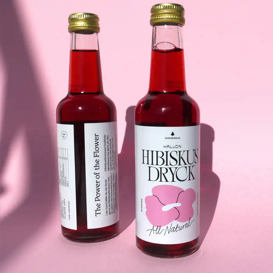 Två flaskor Hibiskus Dryck med hallonsmak står mot en rosa bakgrund. Den ena flaskan visar framsidan av etiketten med en stilren design och en rosa hibiskusblomma, medan den andra flaskan visar baksidan med texten "The Power of the Flower" och produktinformation. Dryckens djupröda färg reflekteras i ljuset och skapar skuggor på bakgrunden.