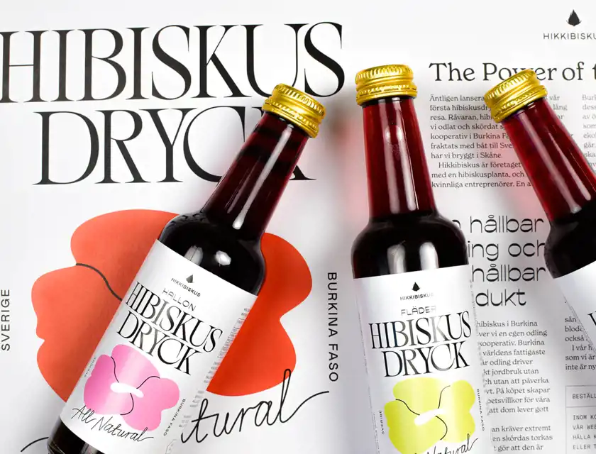 Tre flaskor Hibiskus Dryck med smakerna hallon och fläder ligger på en bakgrund med text och grafik som beskriver drycken. Etiketterna har en stilren design med blommotiv i rosa och grönt. Orden "Hibiskus Dryck" och "The Power of the Flower" syns tydligt på bakgrunden, tillsammans med information om hållbar produktion och ursprung i Burkina Faso.
