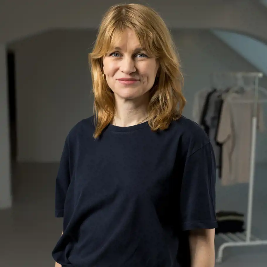 Porträttfoto på Hanna Saurén, medgrundare av varumärket Imperfect Industries. Hon har axellångt blont hår, bär en mörkblå tröja och ler vänligt mot kameran. I bakgrunden syns en ljus, minimalistisk inomhusmiljö med klädhängare.