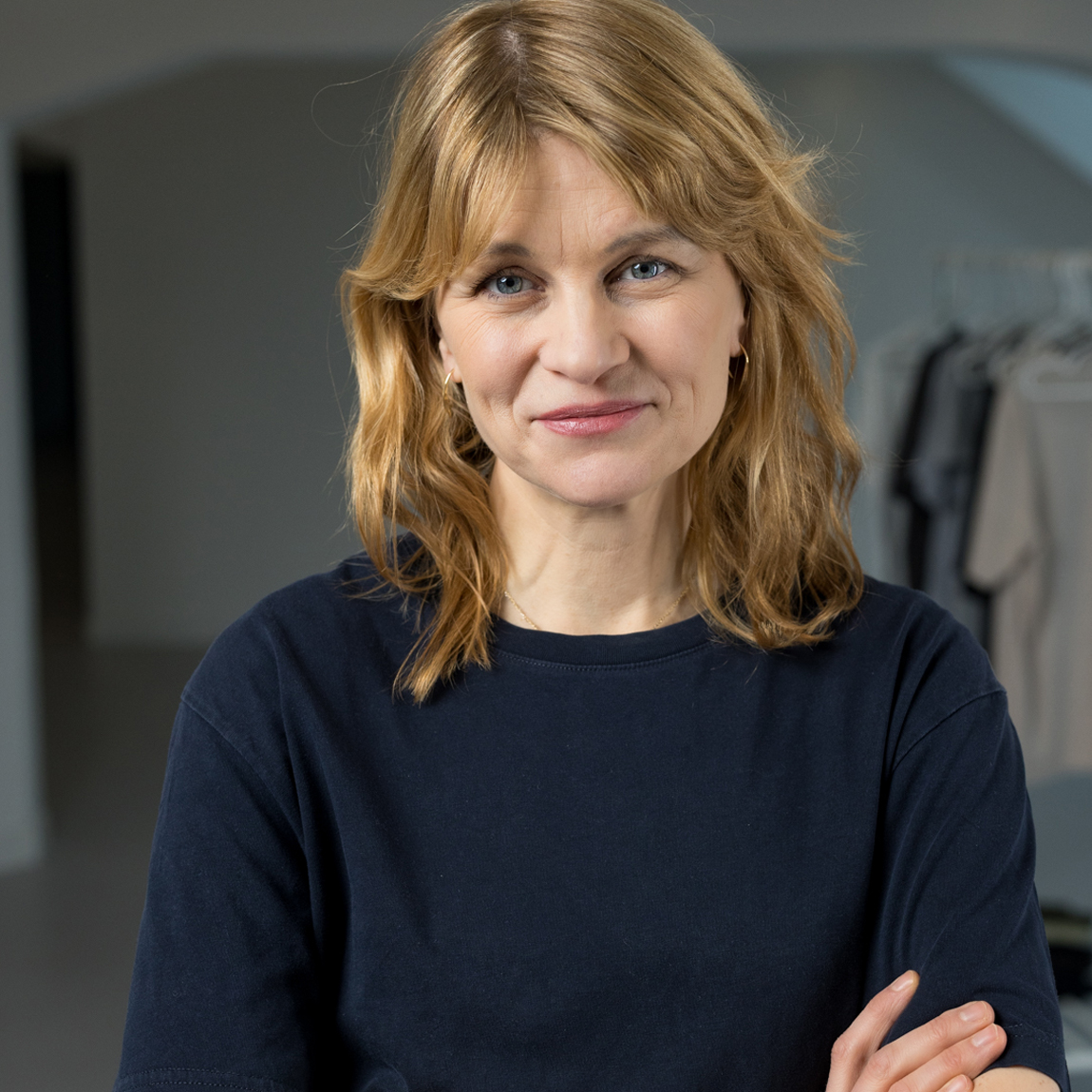 Porträttfoto på Hanna Saurén, medgrundare av varumärket Imperfect Industries. Hon har axellångt blont hår, bär en mörkblå tröja och ler vänligt mot kameran. I bakgrunden syns en ljus, minimalistisk inomhusmiljö med klädhängare.