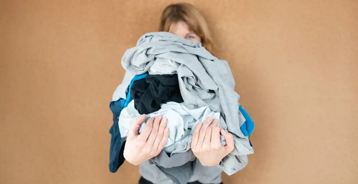 Hanna Saurén, medgrundare av Imperfect Industries, står mot en beige bakgrund och håller en stor hög med vikta eller skrynkliga kläder i olika färger framför sig så att nästan hela ansiktet döljs. Fokus ligger på textilerna och hennes händer, vilket skapar ett lekfullt men tankeväckande uttryck.
