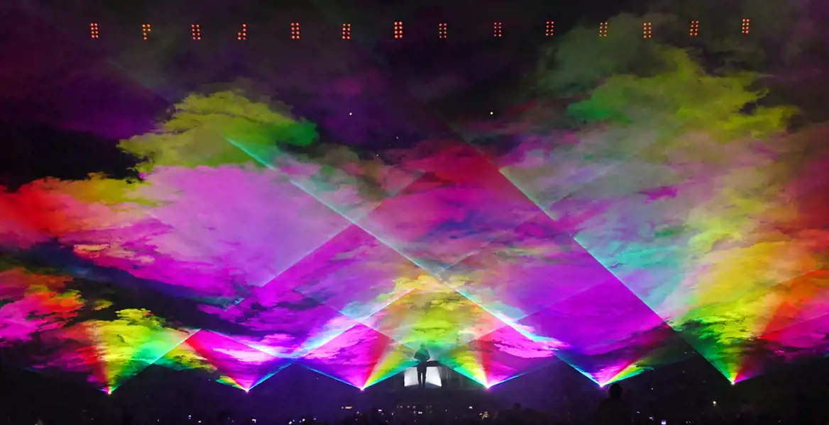 En spektakulär ljusshow med regnbågsfärgade laserstrålar som skapar ett molnliknande mönster över en scen. En silhuett av en DJ syns i mitten, omgiven av intensiva ljuseffekter. Publiken i förgrunden håller upp mobiltelefoner för att filma eller fotografera den färgsprakande upplevelsen. Stämningen är elektrisk och futuristisk.