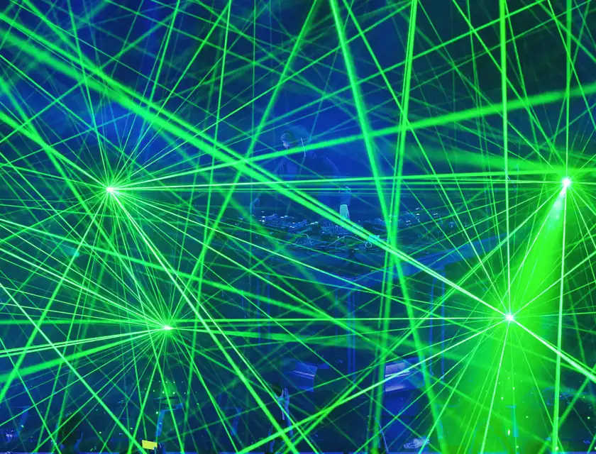 En DJ uppträder på en scen omgiven av en intensiv ljusshow med korsande gröna laserstrålar. Silhuetten av DJ:n är synlig genom ljuseffekterna, medan publiken i förgrunden knappt kan urskiljas genom det bländande skenet. Den elektriska atmosfären och de dynamiska ljusmönstren skapar en futuristisk och energifylld stämning.