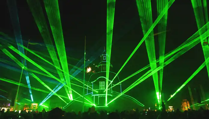 En spektakulär ljusshow med gröna och blå laserstrålar som lyser upp natthimlen. I centrum projiceras konturerna av en historisk byggnad med ett torn i ljusgröna linjer. Publiken syns som mörka silhuetter i förgrunden, medan en fyrverkeriexplosion gnistrar vid sidan av byggnaden. Ljuseffekterna skapar en futuristisk och magisk atmosfär.