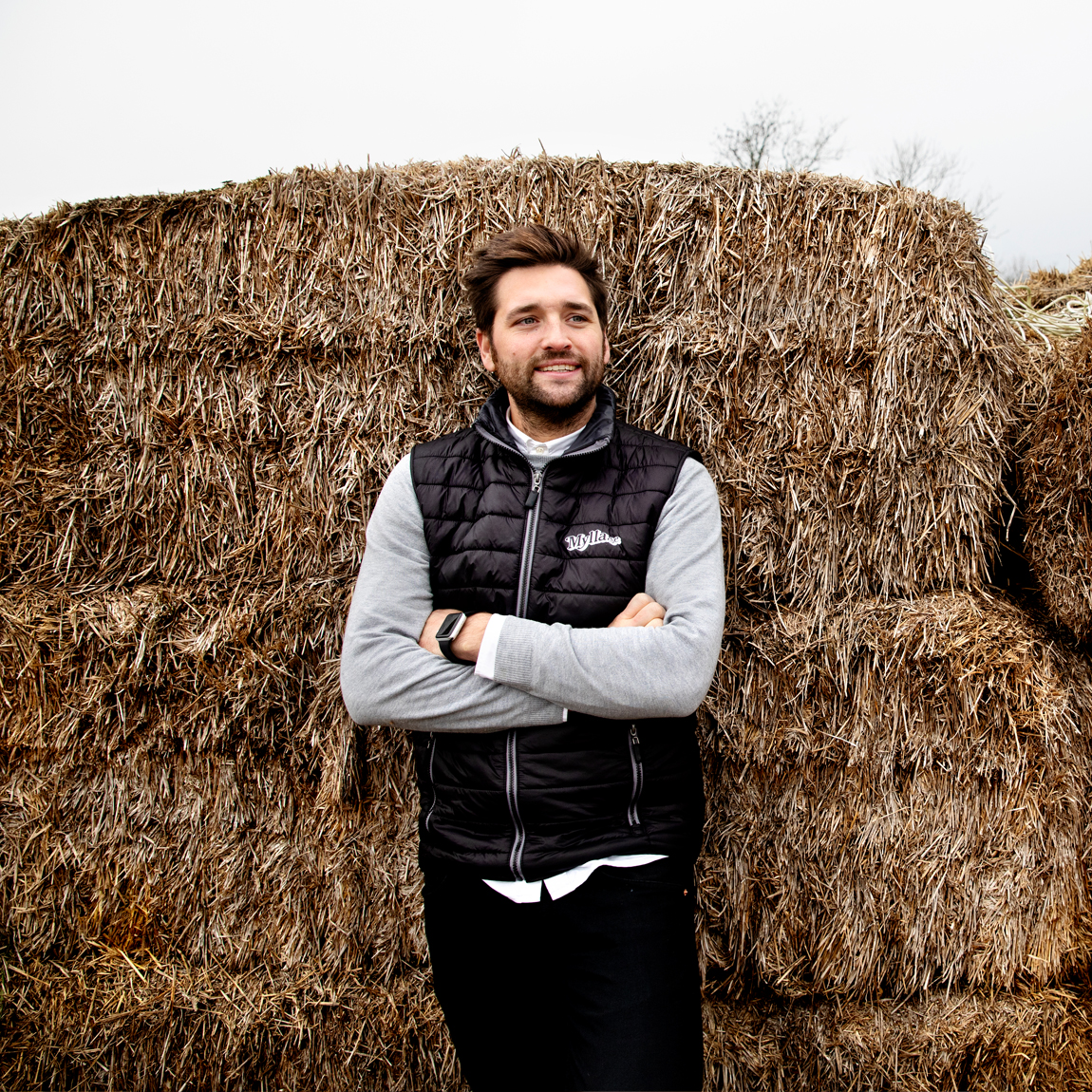 En man med skägg och mörkt hår lutar sig avslappnat mot en stor hög av halmbalar. Han bär en svart vadderad väst med en logotyp på bröstet, en grå långärmad tröja och en smartklocka på handleden. Bakgrunden är en mulen himmel med några trädtoppar i fjärran.