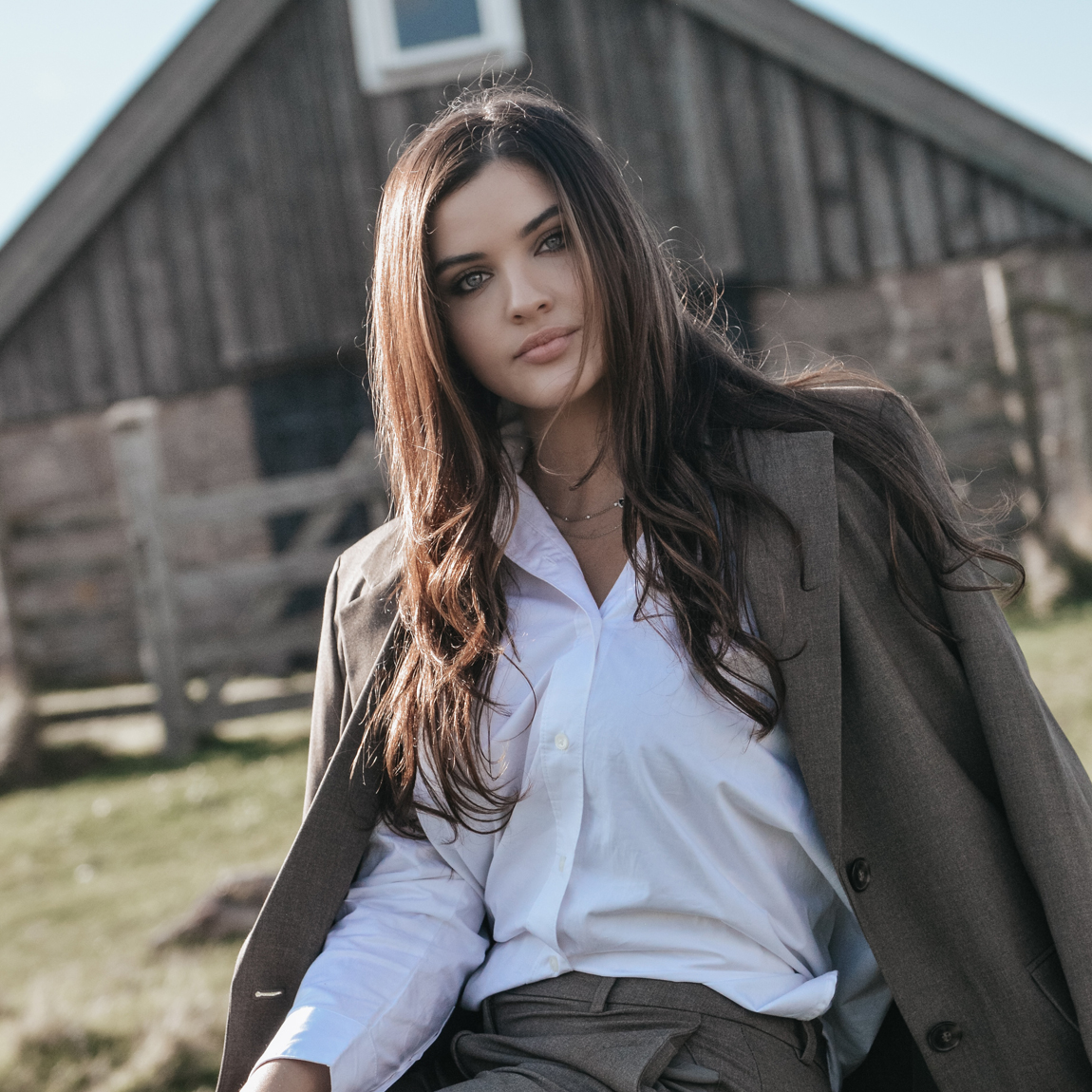 Nadine Alamine, grundare av Myuk, sitter utomhus framför en träbyggnad. Hon bär en vit skjorta och brun kostym och tittar in i kameran i naturligt ljus.