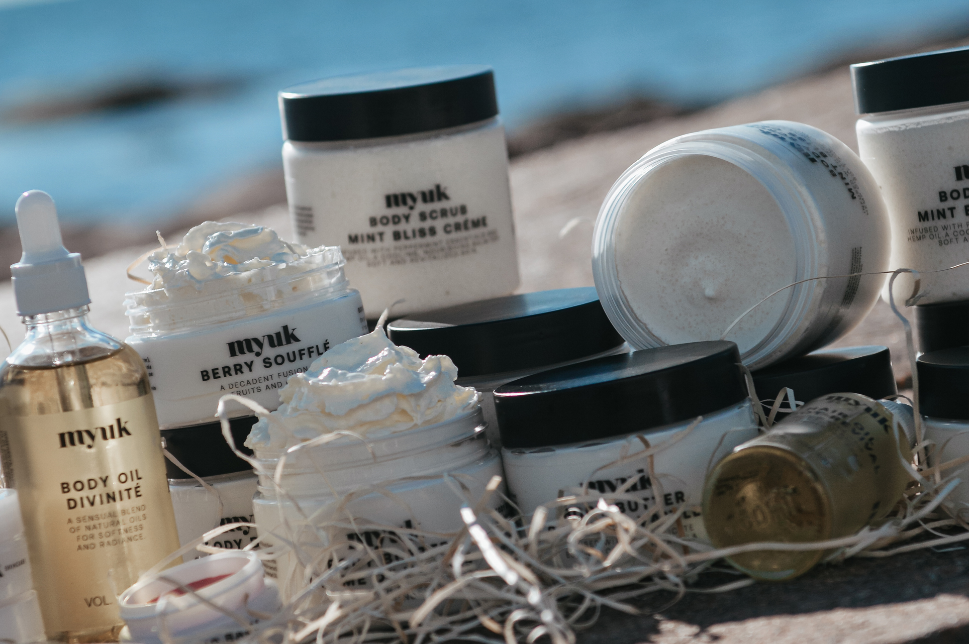 Hudvårdsprodukter från Myuk uppställda på en sten vid havet. Burkar och flaskor med body scrub, body oil och krämer i naturligt ljus.