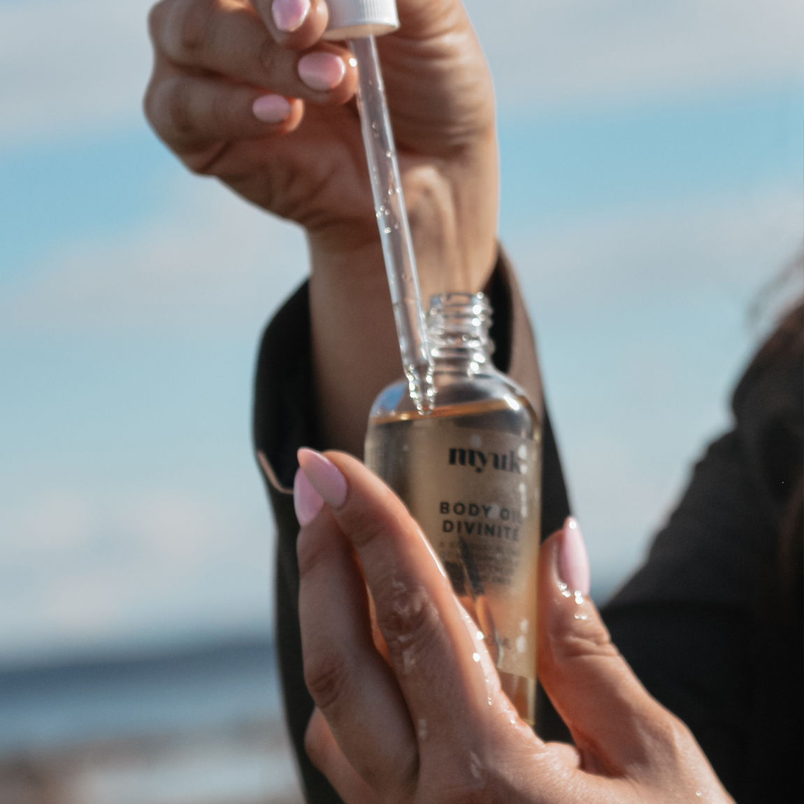 Närbild på händer som håller i en glasflaska med pipett fylld med Myuk Body Oil Divinité, mot en bakgrund av himmel och hav.