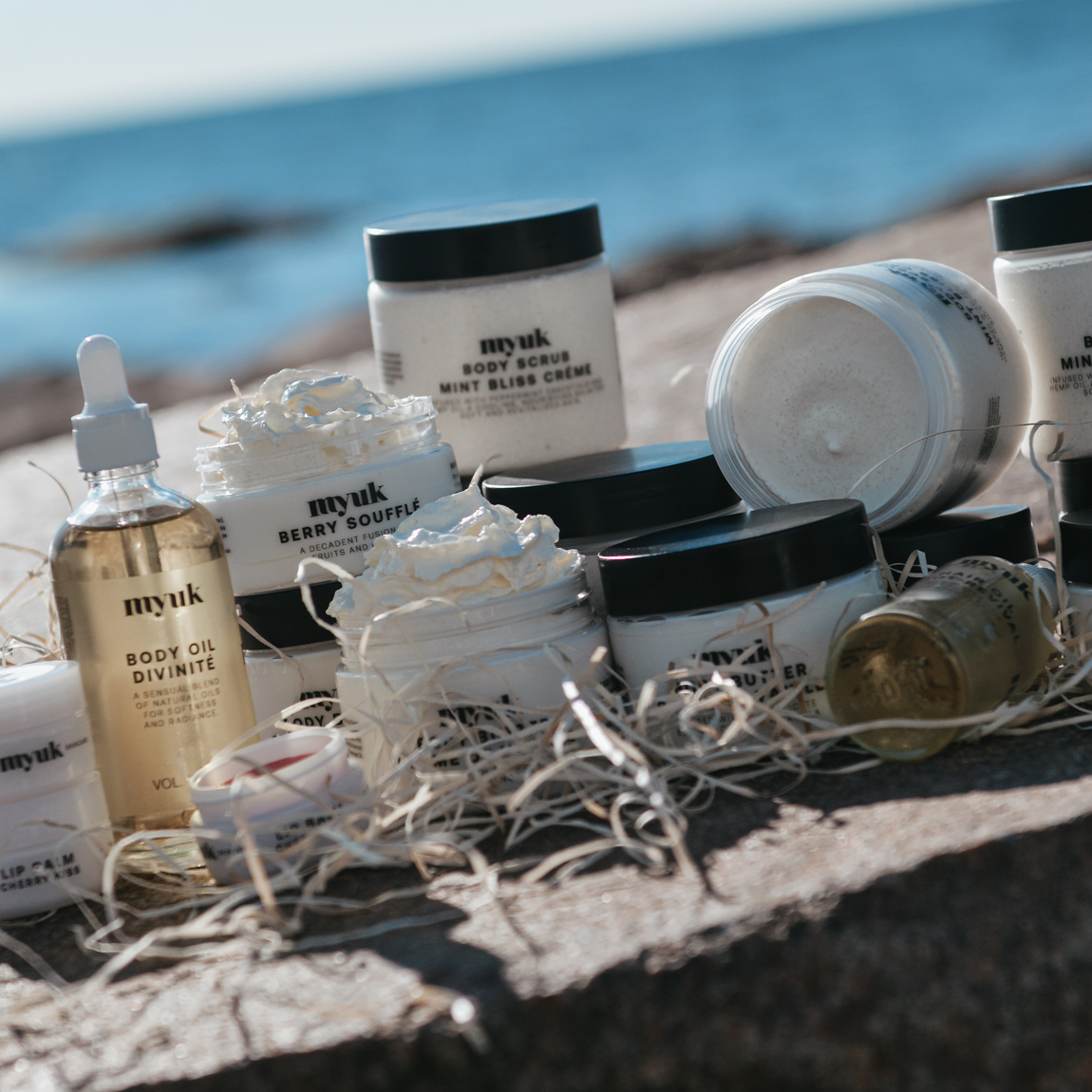 Hudvårdsprodukter från Myuk uppställda på en sten vid havet. Burkar och flaskor med body scrub, body oil och krämer i naturligt ljus.