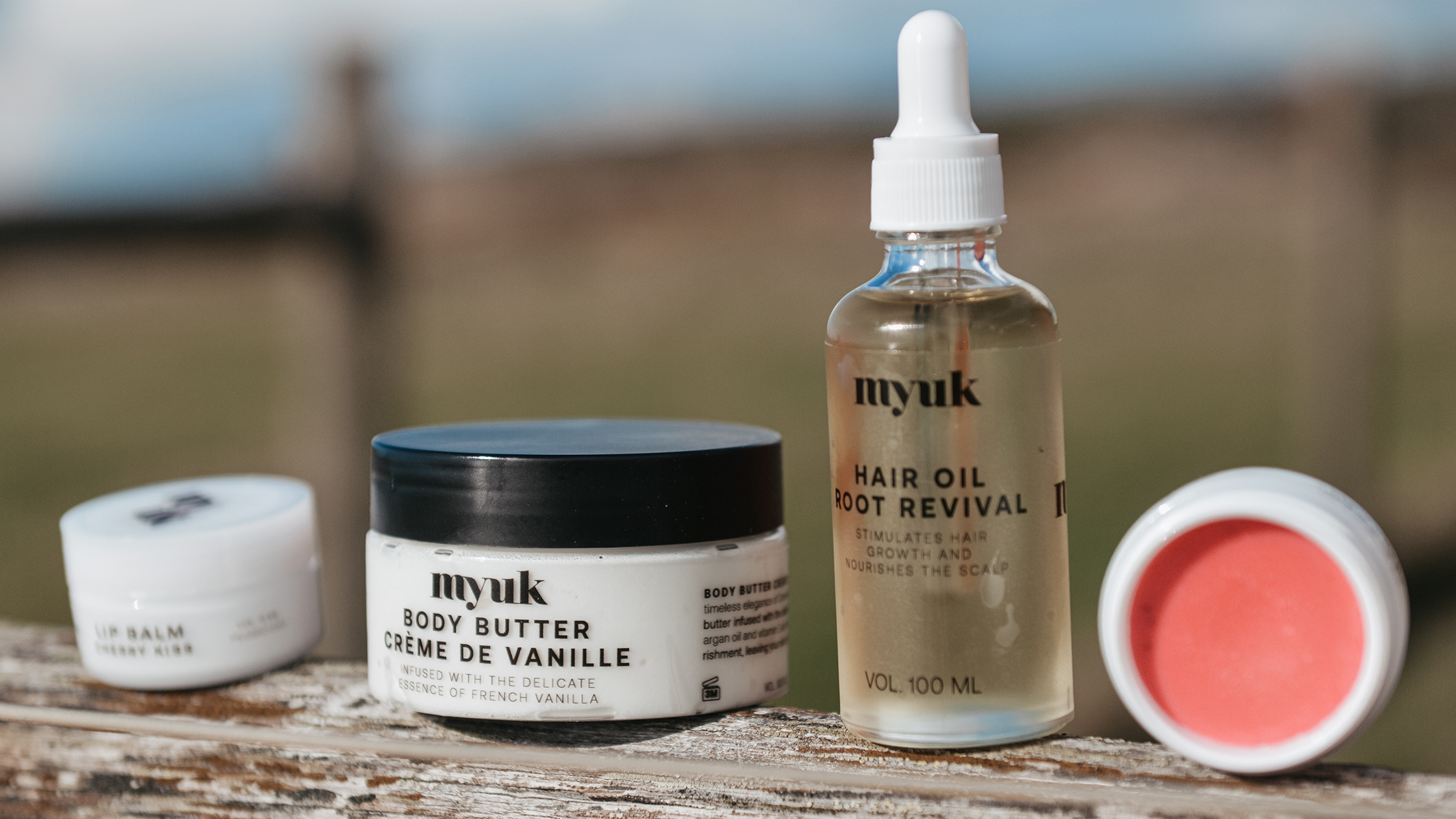 Myuk-produkter uppställda utomhus på en träyta. En burk body butter, en flaska hårolja och två läppbalsam i naturligt ljus med suddig bakgrund.