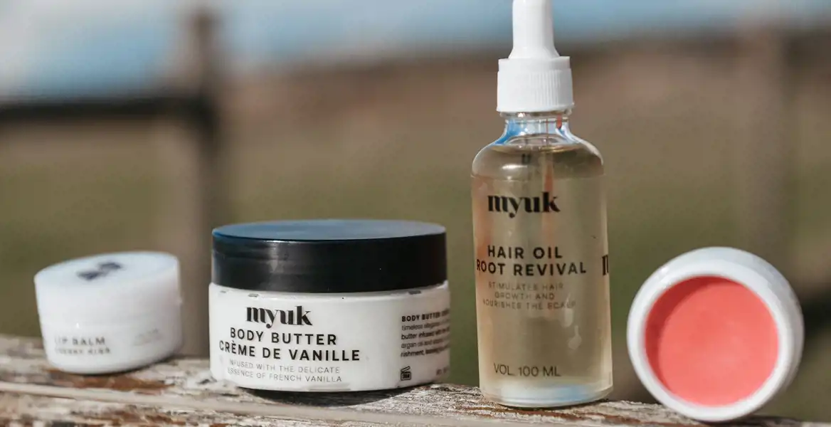 Myuk-produkter uppställda utomhus på en träyta. En burk body butter, en flaska hårolja och två läppbalsam i naturligt ljus med suddig bakgrund.