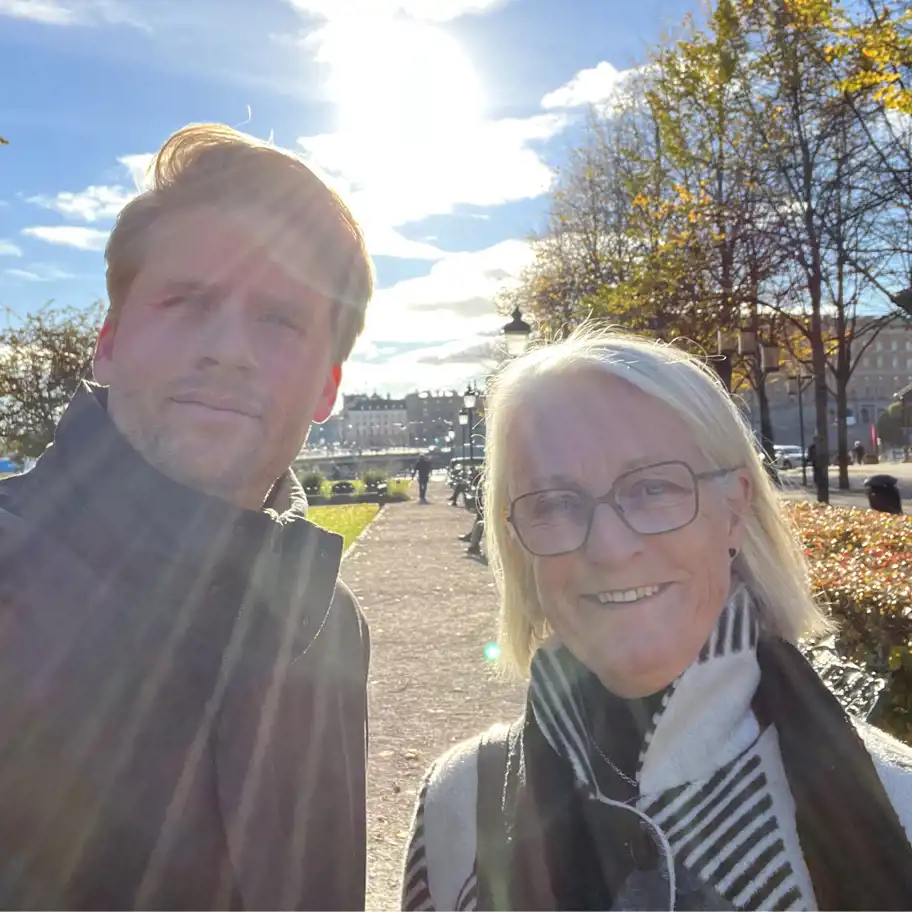 Två personer står utomhus i solskenet, med solstrålar som lyser genom bilden. Den ena personen är en man i en mörk jacka, och den andra är en kvinna med glasögon och en svartvit halsduk. De ler mot kameran med en stadsmiljö i bakgrunden, där träd med höstlöv och en promenadväg syns.