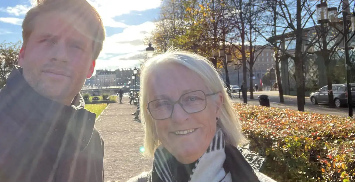 Två personer står utomhus i solskenet, med solstrålar som lyser genom bilden. Den ena personen är en man i en mörk jacka, och den andra är en kvinna med glasögon och en svartvit halsduk. De ler mot kameran med en stadsmiljö i bakgrunden, där träd med höstlöv och en promenadväg syns.